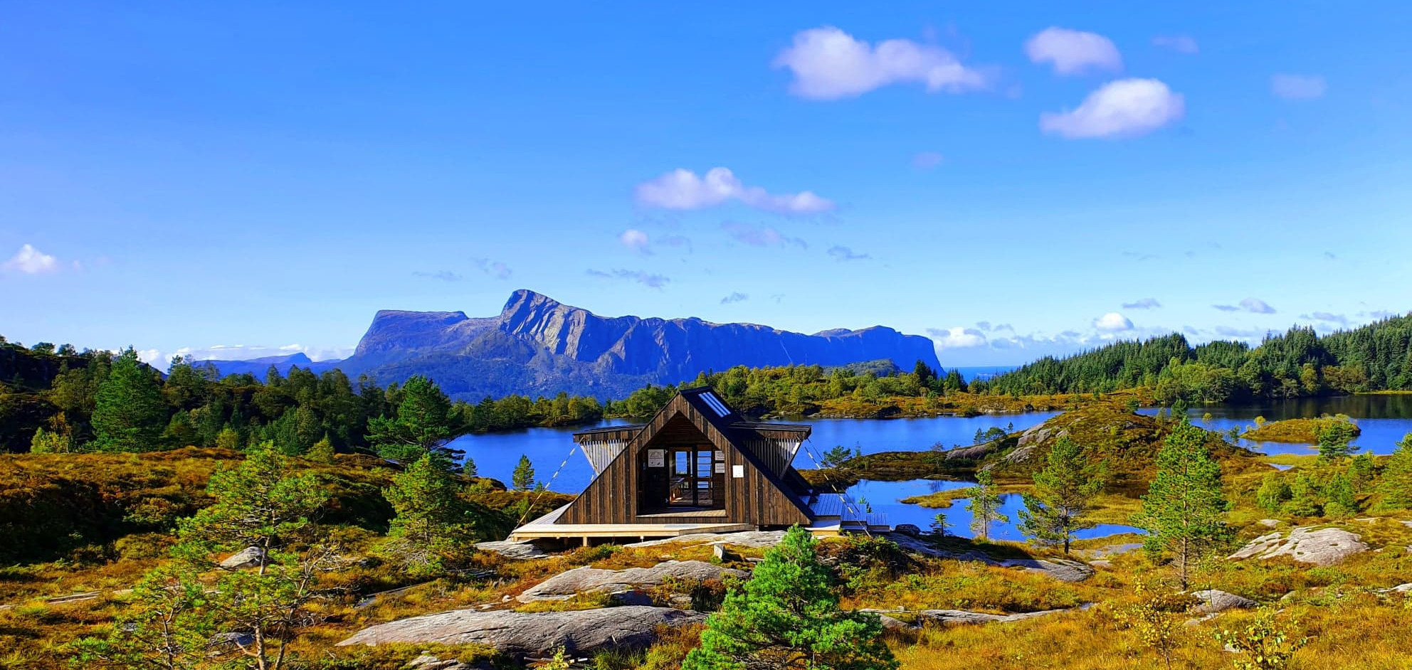Bilete av ei hytte med vatn og fjell i bakgrunnen